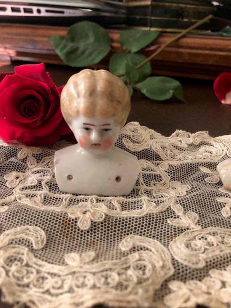 May include: A porcelain doll head with blonde hair and a painted face. The head is on a white lace doily.