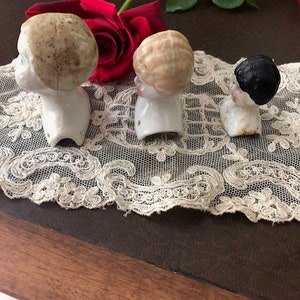 May include: Three vintage porcelain doll heads with different hairstyles. The heads are white with painted features and are missing their bodies. The heads are sitting on a white lace doily.