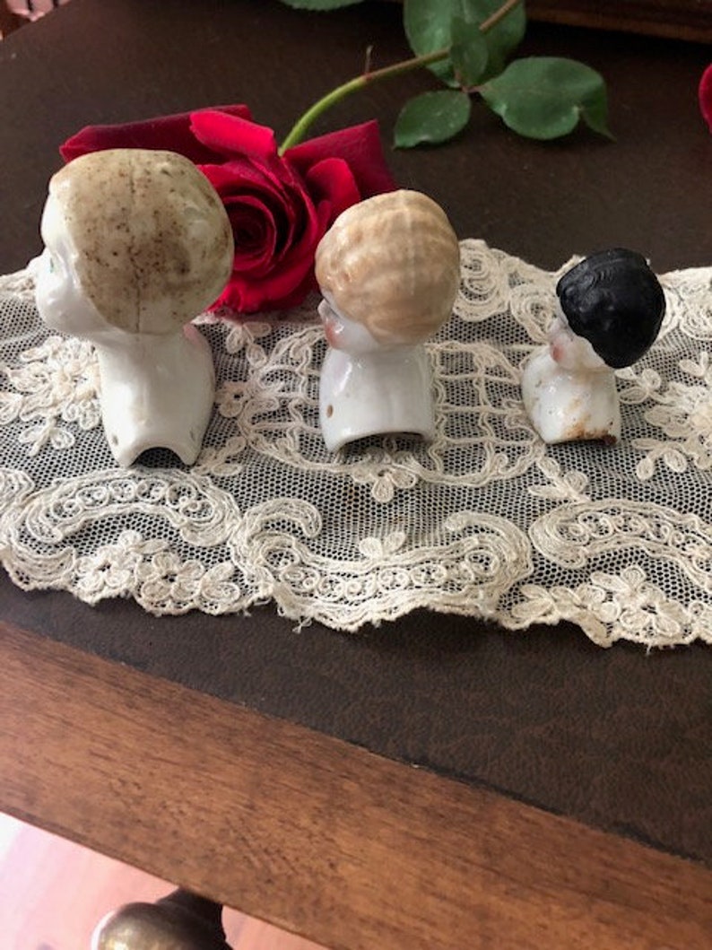 May include: Three vintage porcelain doll heads with different hairstyles. The heads are white with painted features and are missing their bodies. The heads are sitting on a white lace doily.