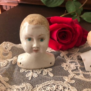 May include: A porcelain doll head with painted blue eyes and a light brown painted hairstyle. The doll head is sitting on a white lace doily with a red rose behind it.