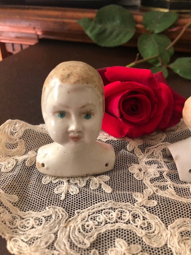 May include: A porcelain doll head with painted blue eyes and a light brown painted hairstyle. The doll head is sitting on a white lace doily with a red rose behind it.
