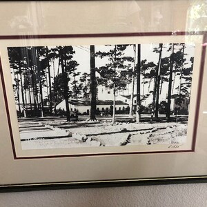 May include: Black and white photograph of a building with a white facade surrounded by trees. The building has a gabled roof and several windows. The trees are tall and slender, with their branches reaching up towards the sky. The photograph is framed in a black frame with a red mat.