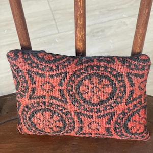 May include: A small, rectangular pillow with a woven design in red and black. The pillow features a repeating circular pattern and is resting on a wooden chair.