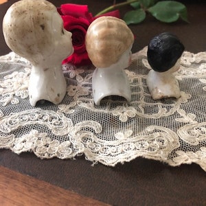 May include: Three porcelain doll heads with different hair colors and styles. The heads are on a white lace doily. The doll heads are vintage and have a delicate, antique look.