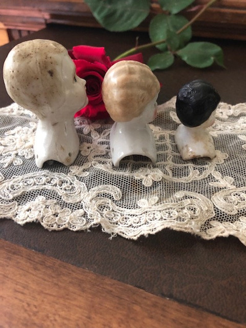 May include: Three porcelain doll heads with different hair colors and styles. The heads are on a white lace doily. The doll heads are vintage and have a delicate, antique look.