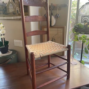 May include: A wooden ladder-back chair with a woven seat. The chair has a dark reddish-brown frame and a light-colored woven seat. The chair is placed on a dark wooden table.