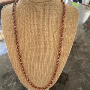 May include: A copper-colored chain necklace displayed on a mannequin. The necklace is made of small, interconnected links, creating a rope-like appearance. The warm, reddish-brown hue of the metal contrasts with the neutral background.