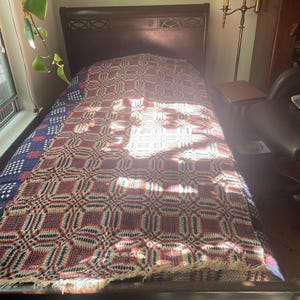May include: A rectangular woven coverlet featuring a repeating geometric pattern in shades of red, navy blue, and cream. The textile has a textured surface with a fringed edge at the bottom, draped over a dark wood bed frame with a headboard.