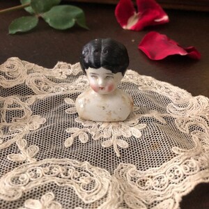 May include: A vintage porcelain doll head with black hair and painted features. The doll head is sitting on a white lace doily.