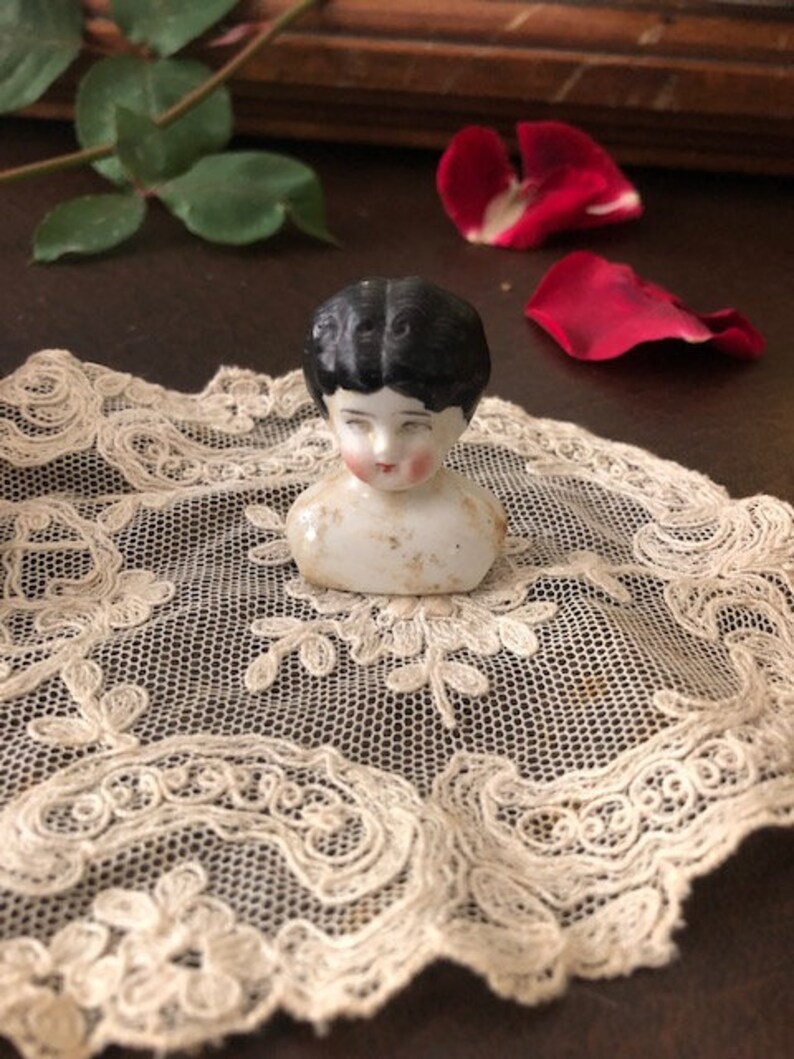 May include: A vintage porcelain doll head with black hair and painted features. The doll head is sitting on a white lace doily.