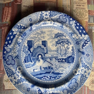 May include: Blue and white decorative plate with a landscape scene. The plate features a detailed illustration of a bridge, trees, and buildings. The rim of the plate has a floral and scroll design. A classic piece for display or dining.