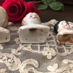 May include: Three porcelain doll heads with painted faces and hair. The heads are attached to white porcelain bases. The heads are sitting on a white lace surface.
