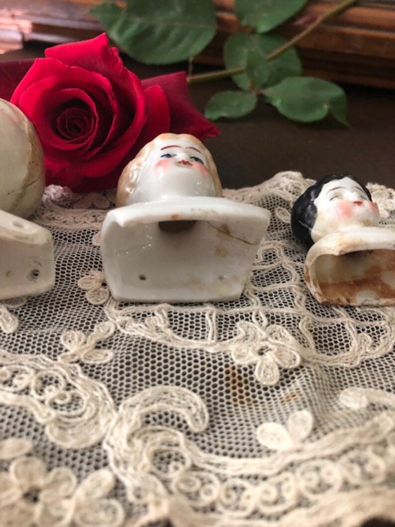 May include: Three porcelain doll heads with painted faces and hair. The heads are attached to white porcelain bases. The heads are sitting on a white lace surface.