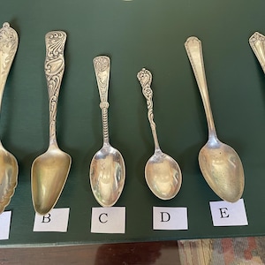 May include: An assortment of antique silverware, including spoons and a fork, arranged on a green surface. The spoons vary in size and design, with ornate handles and bowl shapes. The fork has three tines.