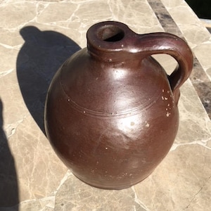 May include: A brown stoneware jug with a handle. The jug has a wide mouth and a rounded body. It is sitting on a stone surface.