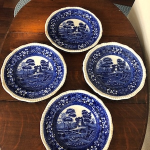 May include: Four blue and white decorative plates with a landscape design. The plates have a scalloped edge and a floral border. The central design depicts a bridge, trees, and a building. The plates are arranged on a dark wooden table.