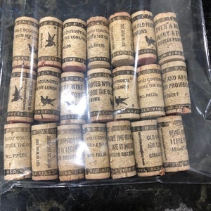 May include: A collection of 15 wine corks with humorous sayings printed on them. The corks are arranged in three rows, with five corks in each row. The sayings on the corks include "What Contented Stole the Cork", "Wine Improves With Age", "But in Wine One Beholds the Heart", "Old Wine Are Our Gods", and "Wine I Like It Any Kind".