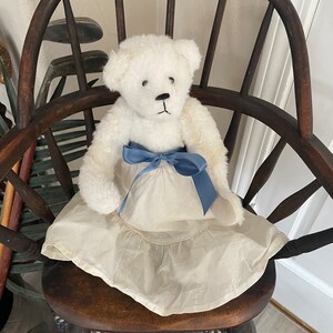 May include: A white teddy bear wearing a white dress with a blue ribbon around the waist, sitting on a wooden chair.