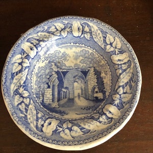 May include: A blue and white ceramic saucer with a floral border and a central image of an arched stone structure.