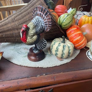 May include: A decorative turkey figurine with a red wattle and brown, black, and white feathers, standing on a round wooden base. It is positioned in front of a woven wicker cornucopia filled with various artificial pumpkins and gourds.
