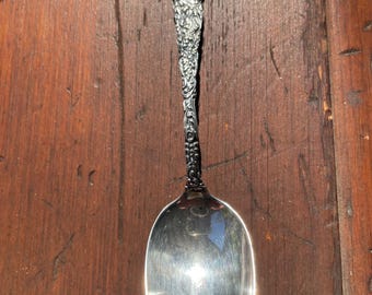 May include: A silver spoon with an ornate floral design on the handle and a heart-shaped detail at the top. The spoon has a polished, reflective bowl. The spoon is likely vintage or antique.