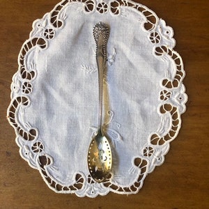 May include: A silver spoon with an ornate handle and a perforated bowl. The spoon is resting on a white lace doily.