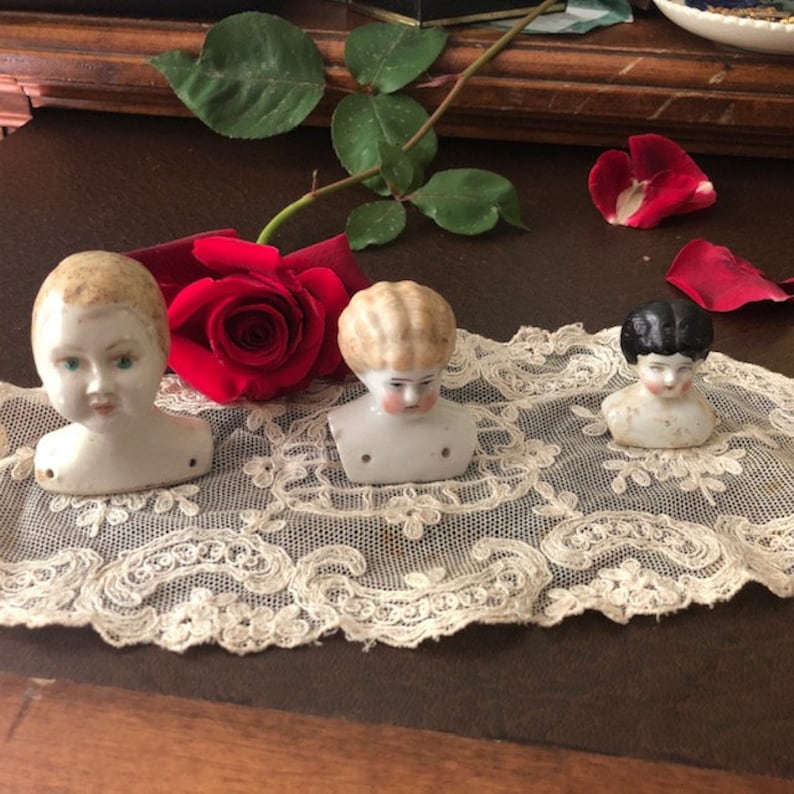 May include: Three porcelain doll heads with painted features. The heads are on a white lace doily. The doll heads are in various shades of white and have painted hair. One doll head has black hair.