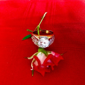 May include: A gold ring with a large, clear diamond set in the center, surrounded by smaller diamonds. The ring is resting on a single red rose bud.