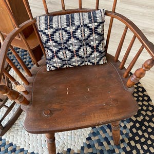 May include: A vintage wooden Windsor chair with a woven pillow. The chair has a curved back with spindles and a rounded seat. The pillow features a geometric pattern in navy, white, and pink. The chair and pillow create a cozy, rustic aesthetic.