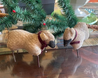May include: Two handcrafted sheep figurines with tan bodies, black faces, and burgundy collars. Each sheep has metal legs and a small bell. A green Christmas tree with red berries is in the background, suggesting a holiday theme.