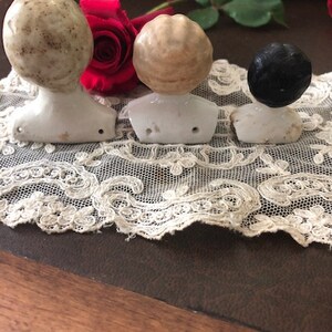 May include: Three porcelain doll heads with different hair colors and styles. The heads are on white porcelain bases and are arranged in a row on a white lace doily.