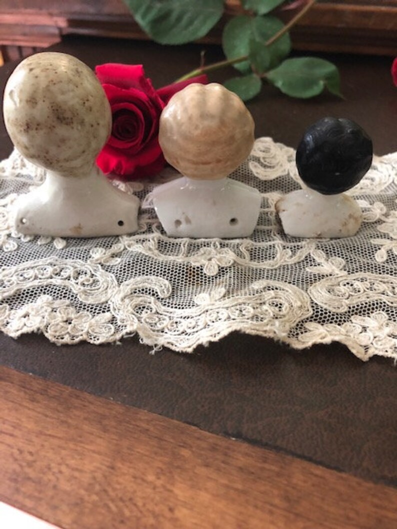 May include: Three porcelain doll heads with different hair colors and styles. The heads are on white porcelain bases and are arranged in a row on a white lace doily.