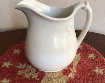 May include: A white ceramic pitcher with a rounded body and a handle. The pitcher has a smooth, glossy finish.