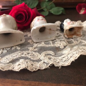 May include: Three porcelain doll heads with painted faces and hair. The heads are attached to white ceramic bases with open bottoms. The heads are arranged on a white lace doily.