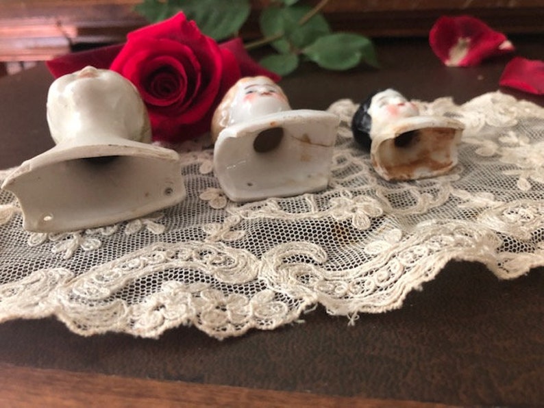 May include: Three porcelain doll heads with painted faces and hair. The heads are attached to white ceramic bases with open bottoms. The heads are arranged on a white lace doily.