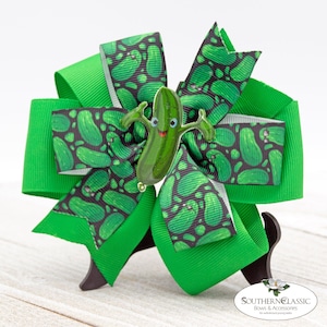 May include: A large, bright green hair bow with a cartoon pickle embellishment. The bow features layers of solid green and patterned ribbon with pickle designs. The pickle charm has a smiling face and outstretched arms. The bow is on a stand.