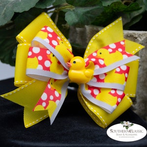 May include: A large, yellow hair bow with a rubber ducky embellishment. The bow features a repeating pattern of yellow ducks and red polka dots on a white background. The bow is accented with gray ribbon and white stitching.