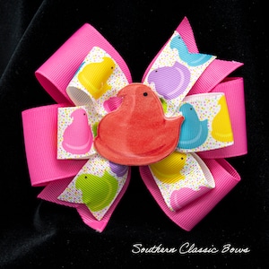 May include: A bright pink hair bow with a red Peep-shaped embellishment. The bow features layers of pink grosgrain ribbon and a patterned ribbon with colorful Peep chicks. The bow is set against a black background.