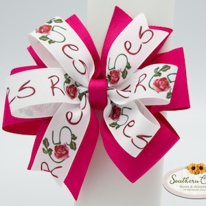 May include: A bright pink hair bow with white ribbon accents. The white ribbon features the word "Rose" in a cursive font, along with a rose illustration. The bow is a layered design, perfect for accessorizing. The bow is a hair accessory.