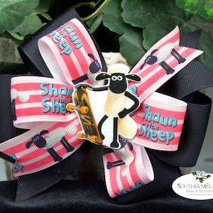 May include: A large hair bow featuring the cartoon character Shaun the Sheep. The bow has black and pink striped ribbons with the text "Shaun the Sheep". A Shaun the Sheep figure with a skateboard is in the center.