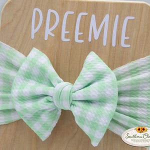 May include: A light green and white gingham baby bow headband. The headband is wrapped around a wooden display board with the word "PREEMIE" in white letters. The bow is centered and has a knotted detail.