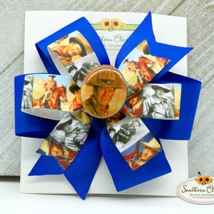 May include: A royal blue hair bow featuring a central image of a man in a cowboy hat, surrounded by smaller images of the same man in various poses. The bow has a layered design with a white backing and the text "Southern Classic Bows & Accessories".