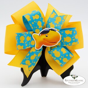 May include: A handmade hair bow featuring layers of yellow and turquoise ribbon. The turquoise ribbon is patterned with yellow goldfish shapes. A cartoon goldfish wearing sunglasses is centered on the bow. The bow is displayed on a black stand.
