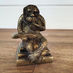 May include: A bronze-colored monkey figurine, seated and reading a book. The monkey is detailed with textured fur and facial features. The figurine is on a small, tiered base, and the overall design suggests a vintage or antique aesthetic.