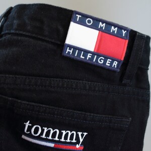 Puede incluir: Primer plano de unos vaqueros de mezclilla negros con el parche del logo de Tommy Hilfiger en el bolsillo trasero. El parche presenta el nombre de la marca y un dise&ntilde;o rojo, blanco y azul. La palabra "tommy" est&aacute; bordada en el bolsillo trasero.