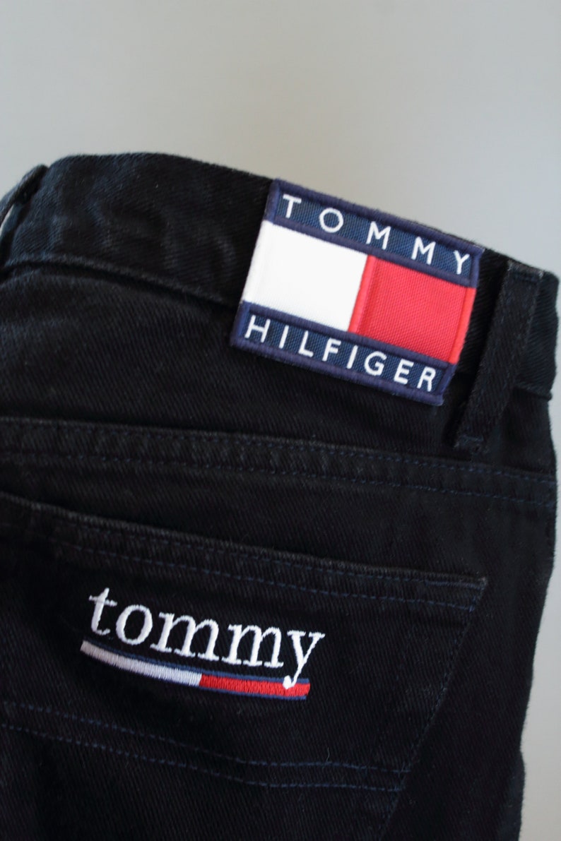 Puede incluir: Primer plano de unos vaqueros de mezclilla negros con el parche del logo de Tommy Hilfiger en el bolsillo trasero. El parche presenta el nombre de la marca y un dise&ntilde;o rojo, blanco y azul. La palabra "tommy" est&aacute; bordada en el bolsillo trasero.