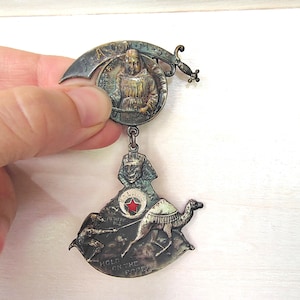May include: Vintage silver-toned metal pin with multiple hanging charms. The top charm features a figure in a circle with text above. Below hangs a charm with a sphinx, a camel, and the words "Hold on the Rope".