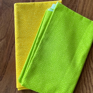 May include: Two folded fabric pieces, one bright yellow and the other vibrant green, both with a repeating square pattern. The fabrics are stacked on a wooden surface, showcasing their texture and color contrast.