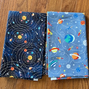 May include: Two folded fabric pieces with space-themed patterns. One features a dark blue background with a solar system design, while the other has a light blue background with planets, rockets, and stars. The fabrics are likely for crafting or sewing projects.