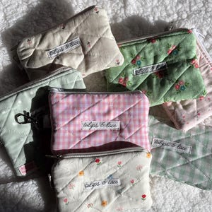 Puede incluir: Una colección de pequeños monederos acolchados en varios colores y estampados. Los monederos presentan estampados florales, cuadros vichy y colores lisos, con un patrón de costura de diamante. Cada monedero tiene cierre de cremallera y una etiqueta que dice "tulips & lace."